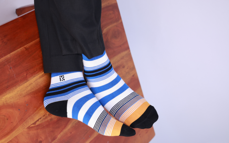 A close-up of a person wearing vibrant striped socks from our bamboo socks collection, paired with black dress trousers. The socks feature a modern pattern of electric blue, black, and white stripes with a distinct yellow accent near the black toe, perfect for adding a pop of color to formal wear.